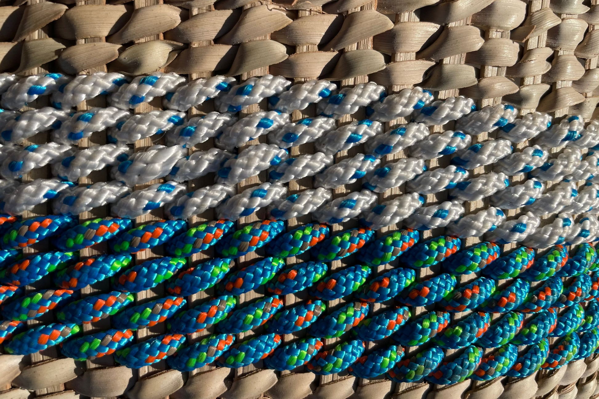 Customisable woven basket with colorful rope. Close-up of braided ropes in white, blue, green, and orange on a woven surface. Ndebele woven wonky basket handmade from natural fibres for home decor, soft furnishings, lodges, restaurants, property developments and interior projects in various customisable sizes, in hues of brown, beige, and peach