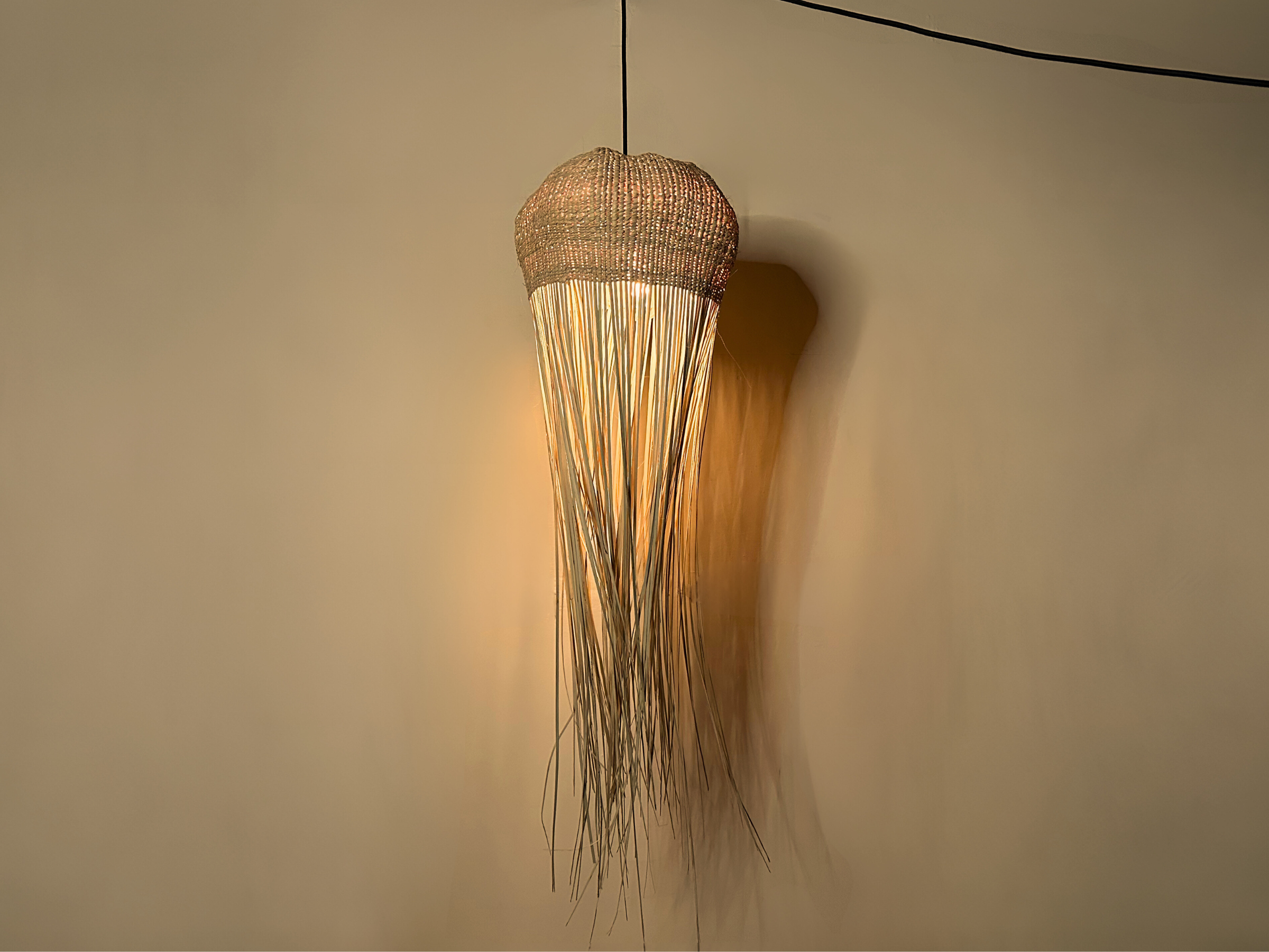 Tall jellyfish-inspired pendant light with woven top and flowing raffia strands, softly glowing against a neutral wall.